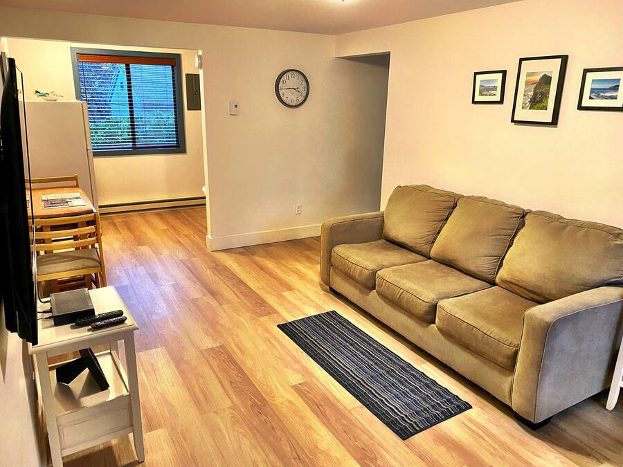 Un salon avec un canapé beige, un parquet en bois, une horloge au mur et une fenêtre avec des stores.