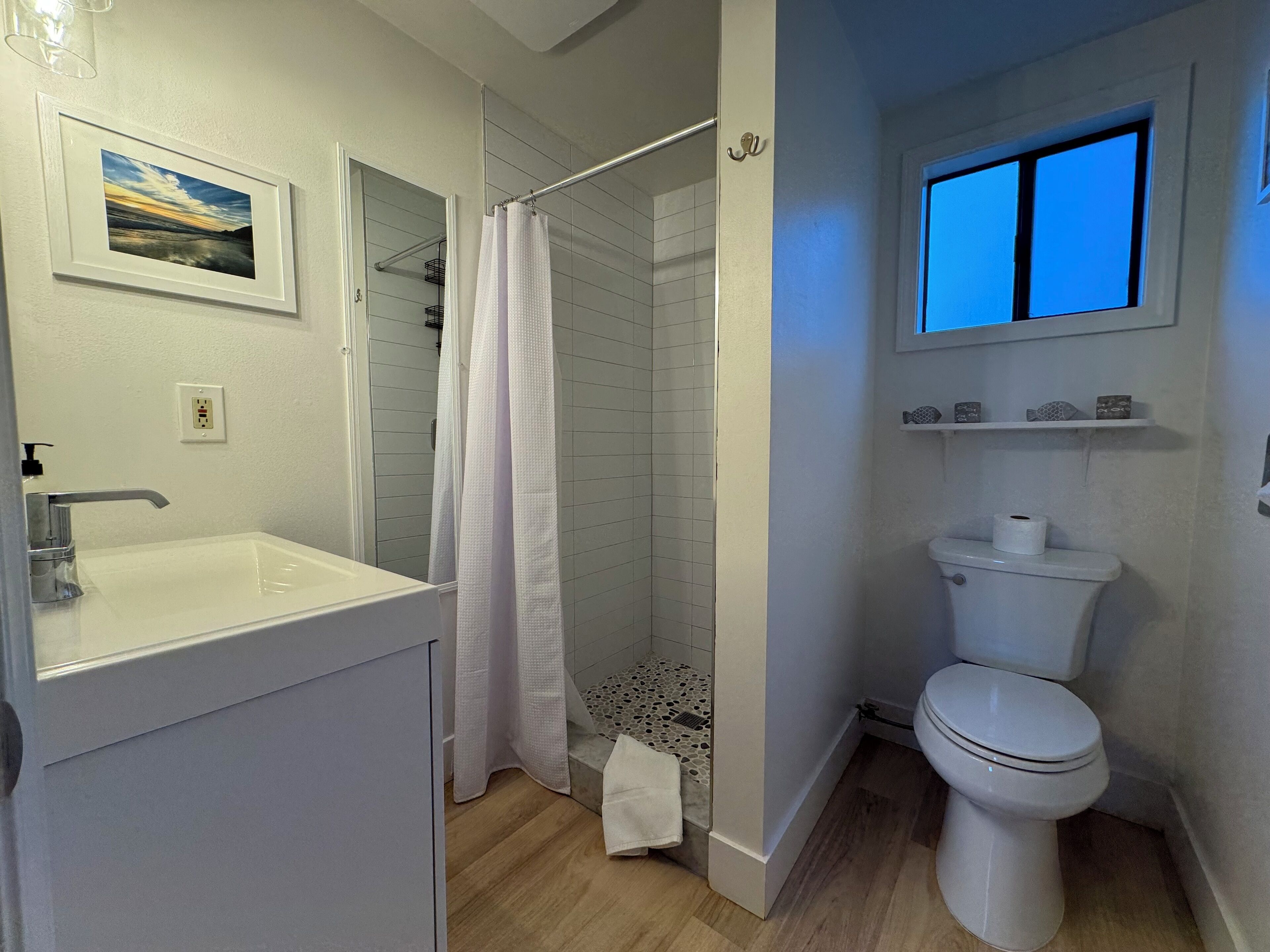 Ein Badezimmer mit einem weißen Waschbecken, einer Toilette und einer Duschkabine. An der Wand hängt ein gerahmtes Bild und über der Toilette befindet sich ein Fenster.