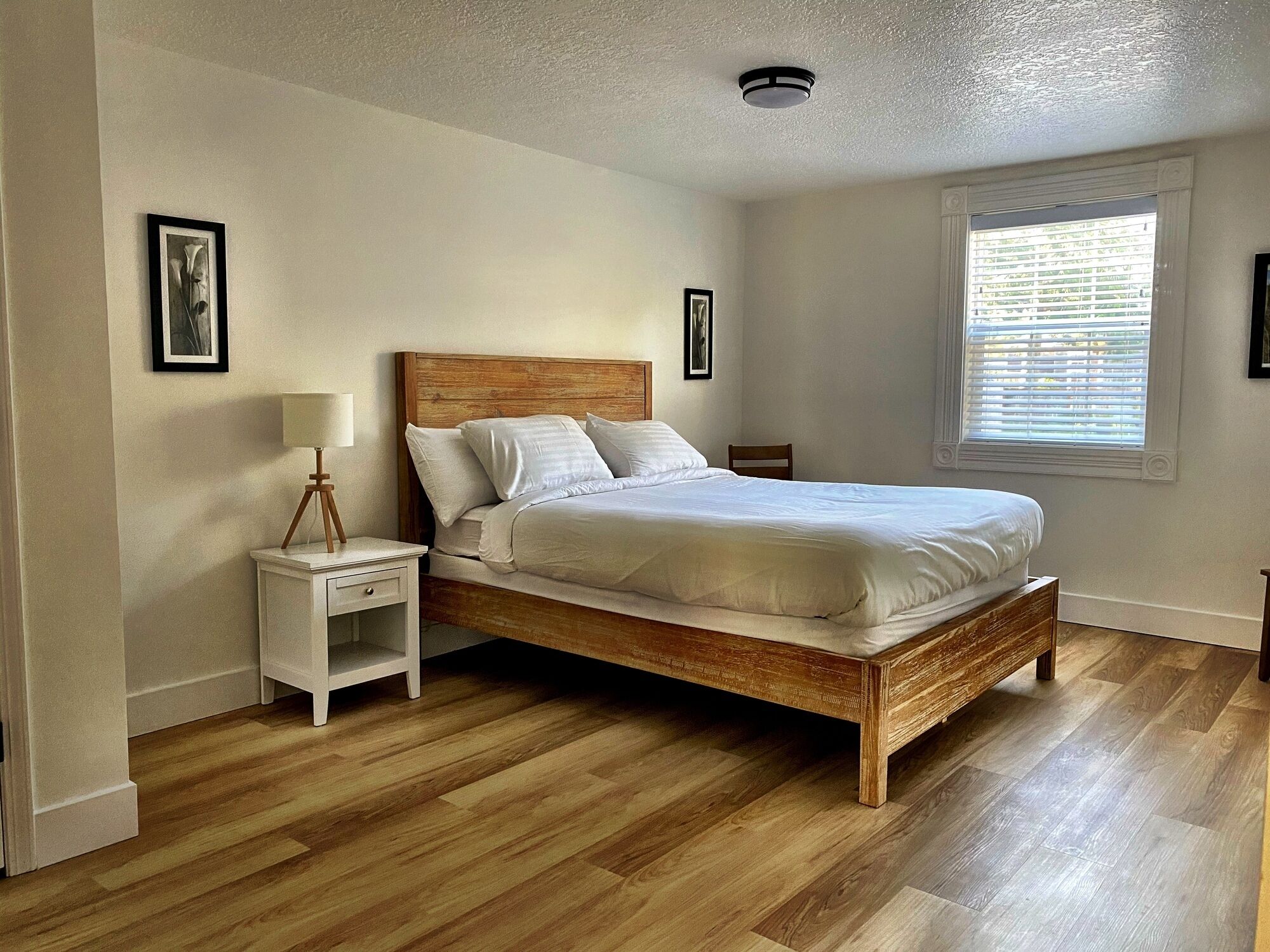 A bedroom with a wooden bed, white bedding, a nightstand with a lamp, and a window with blinds.