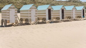 Une rangée de cabanes de plage aux murs rayés et aux toits bleus, chacune dotée d’une porte et d’une roue fixée à l’avant.