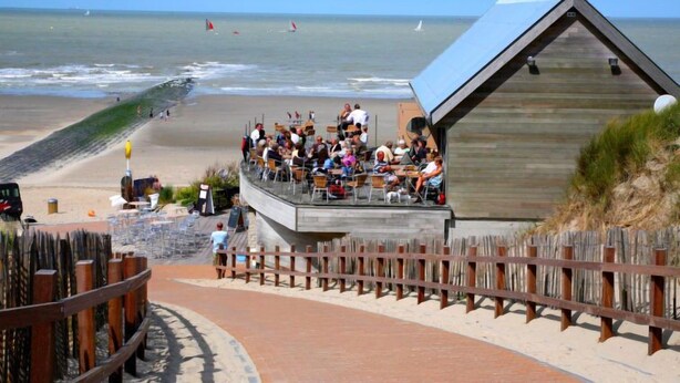 Un restaurant situé en bord de mer, avec une terrasse offrant une vue sur l’océan, un bâtiment en bois et une plage de sable.