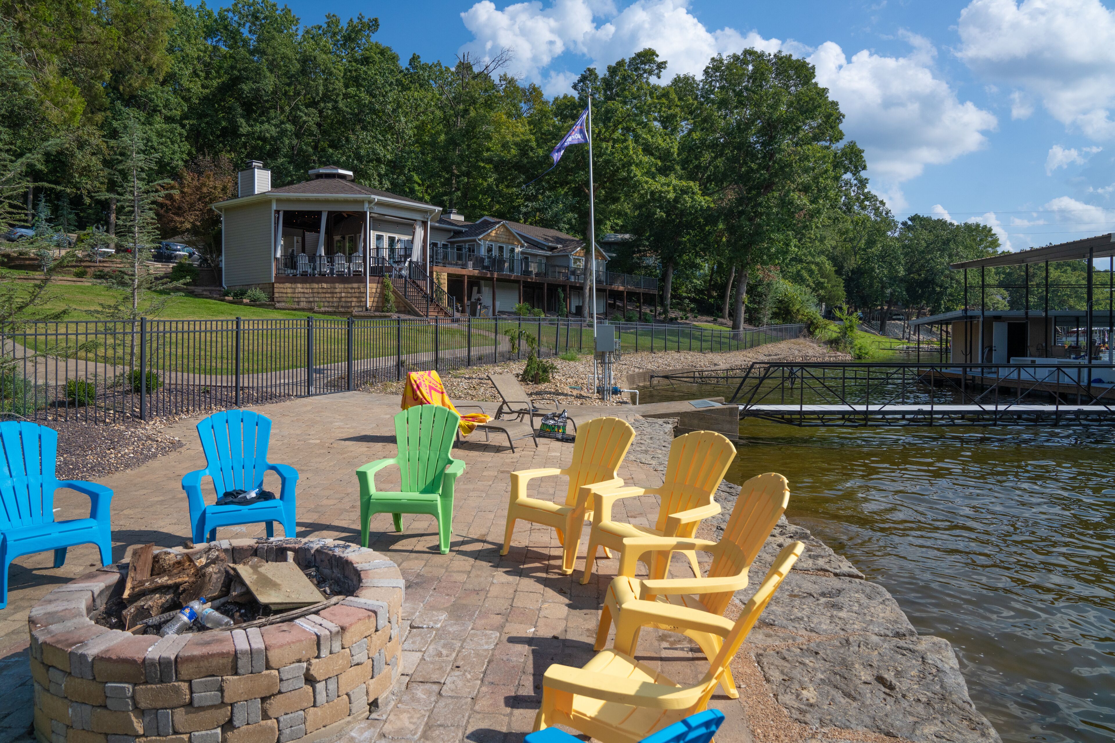 Un patio junto al lago con sillas Adirondack, una chimenea y una casa al fondo.