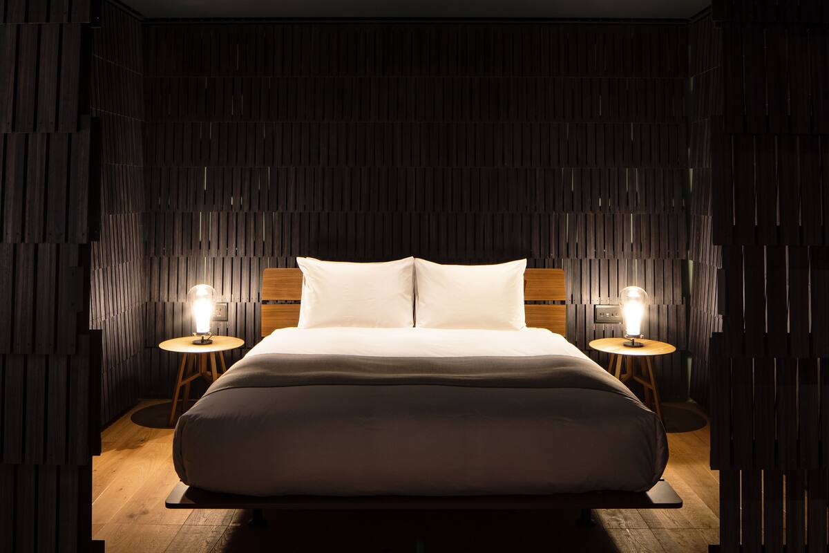A modern bedroom with a wooden headboard, a large bed with white linens, two bedside tables with lamps, and a dark, textured wall.