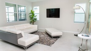A modern living room with a grey sectional sofa, a white ottoman, a small white table, and a floor lamp. - Magical Home in Orlando/Disney/Close to Parks (Kissimmee)