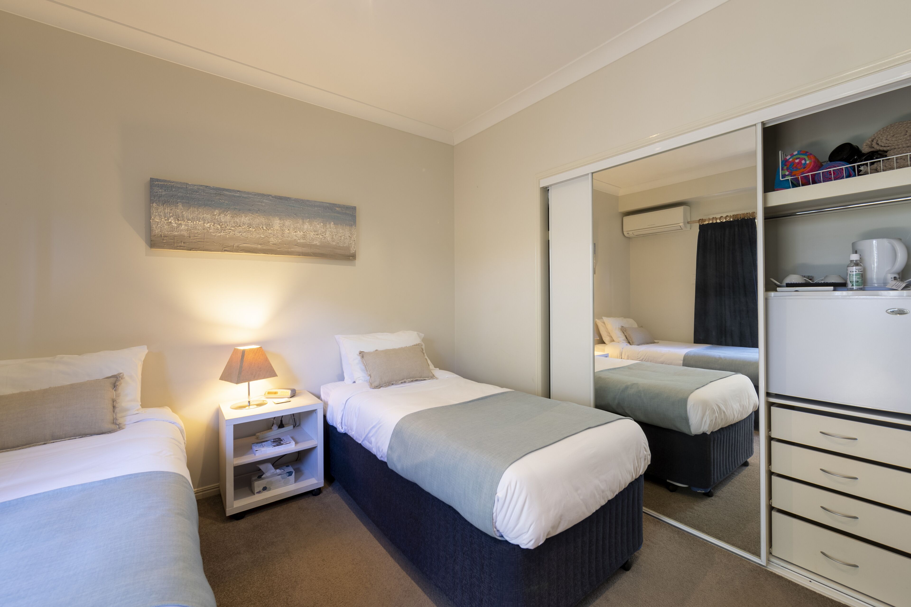 A hotel room with two beds, a bedside table with a lamp, a wardrobe, and a mirror.