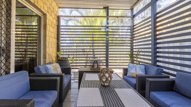 Un espacio de descanso al aire libre con mobiliario de mimbre y una mesa, con vista a palmeras a través de una valla decorativa.