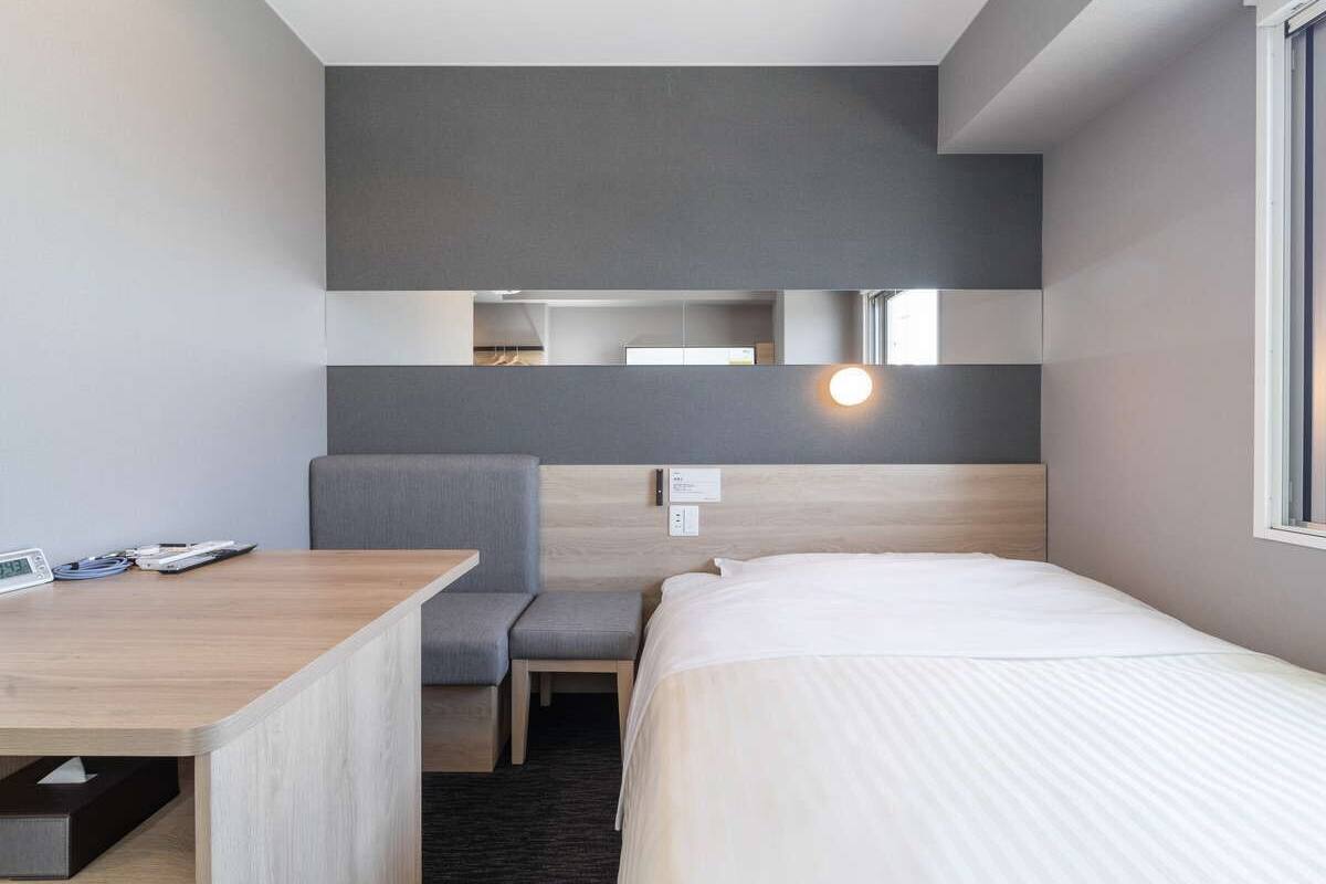 Quarto de hotel com uma cama, uma escrivaninha, uma cadeira e uma mesinha.