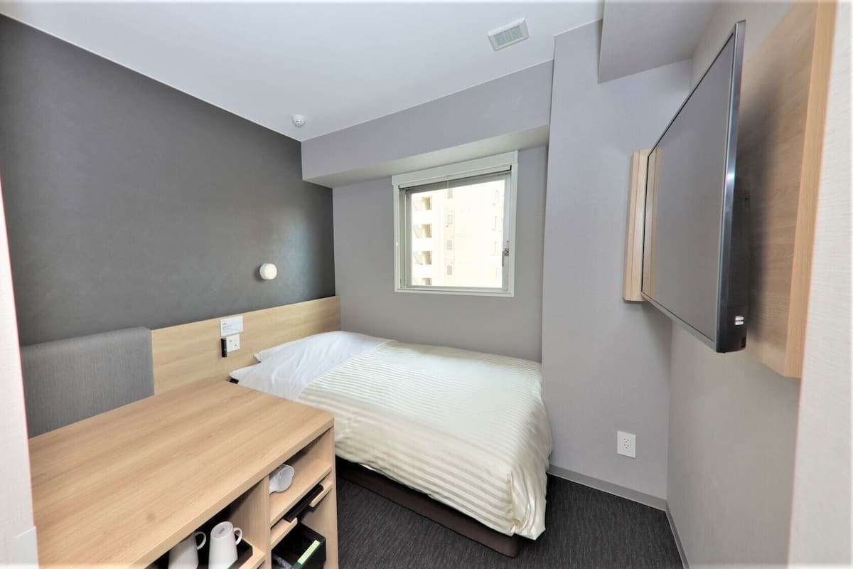 Habitación de hotel pequeña con una cama individual, un escritorio, una silla, una mesita y televisión, además de una ventana.