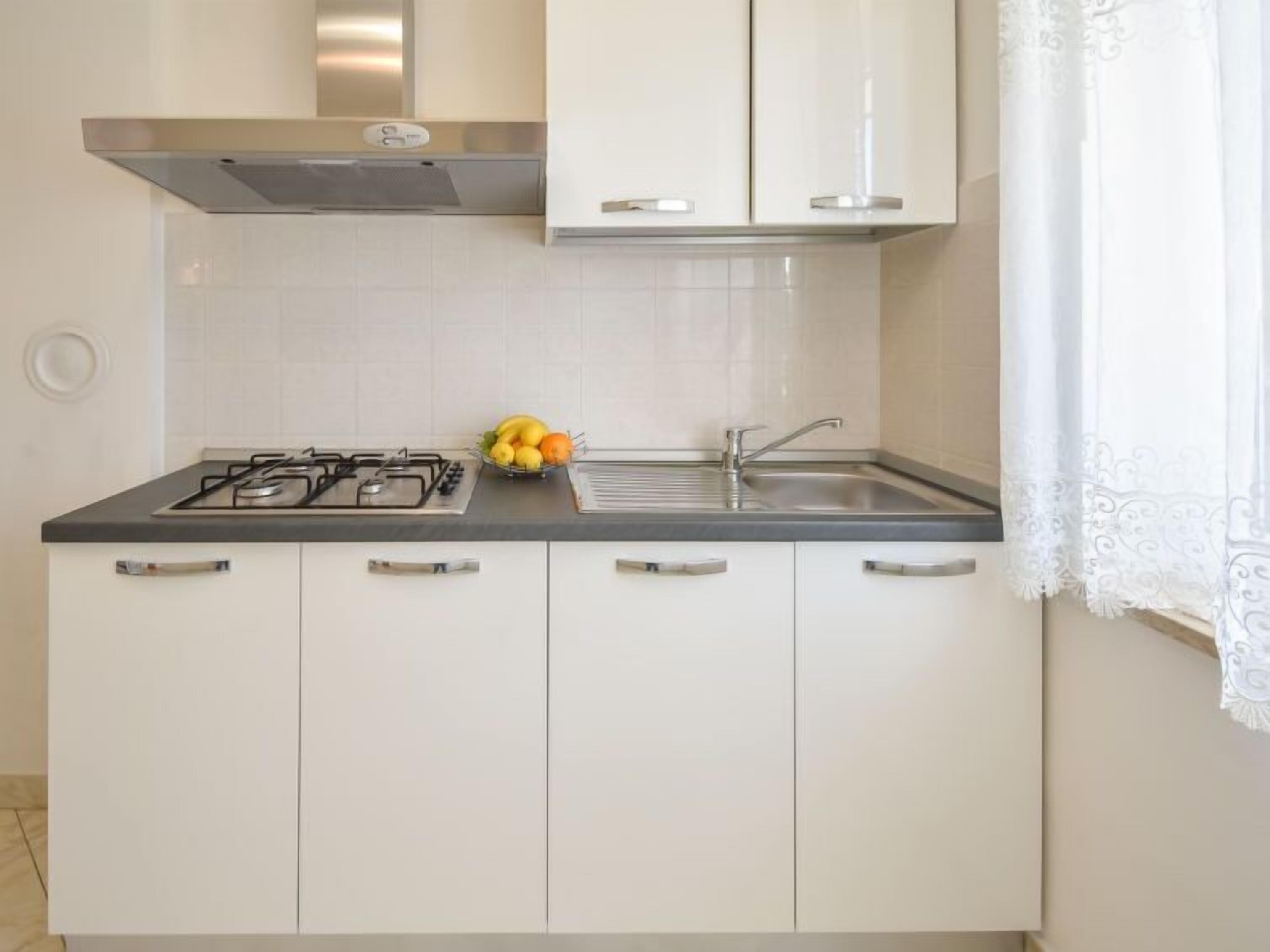 Une cuisine moderne avec des meubles blancs, une cuisinière à gaz, un évier et un panier de fruits posé sur le plan de travail.