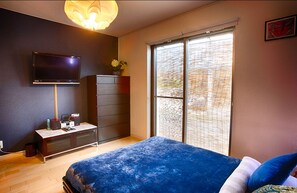 Un dormitorio con cama, televisor montado en la pared, cómoda y ventana con persianas.