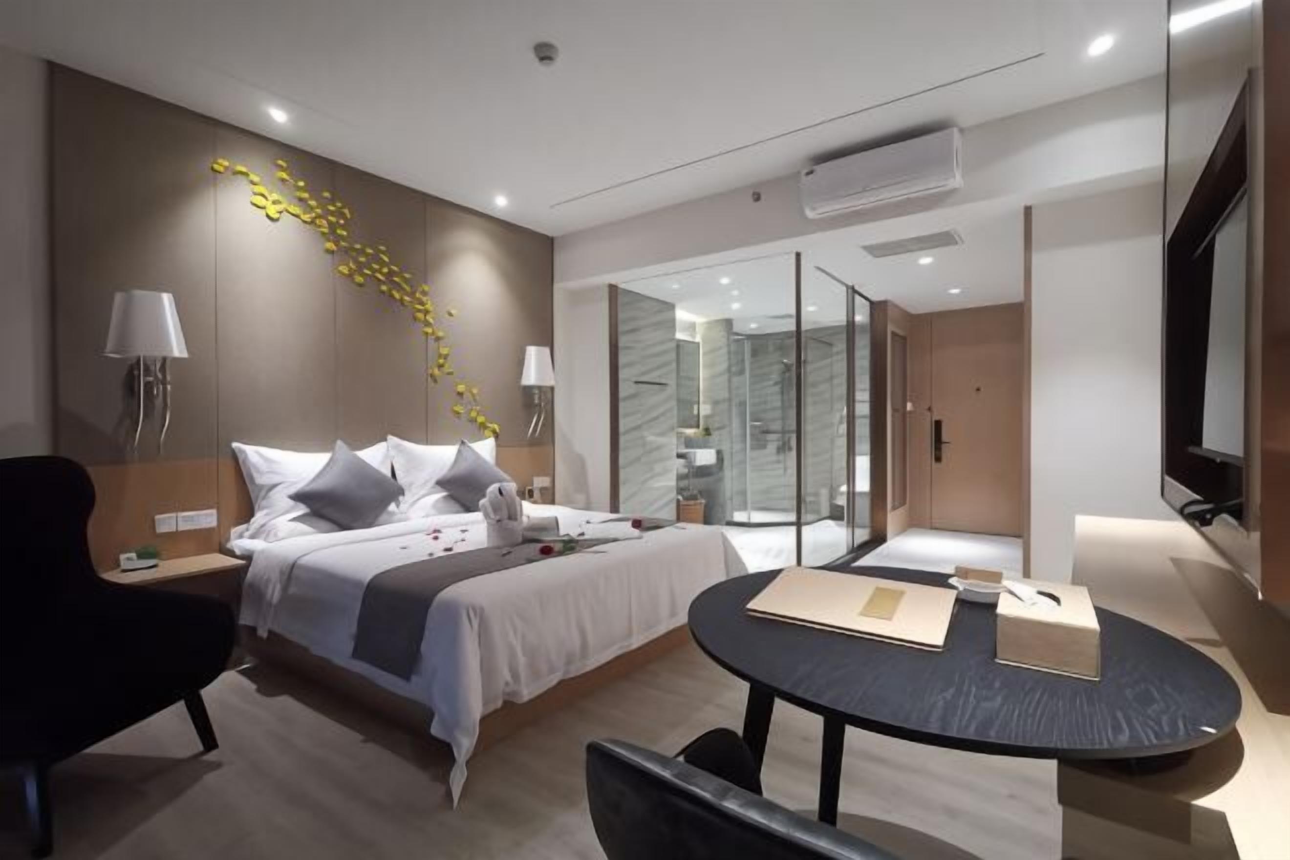 A modern hotel room with a bed, bedside tables, a desk, a chair, a glass shower, and a TV.