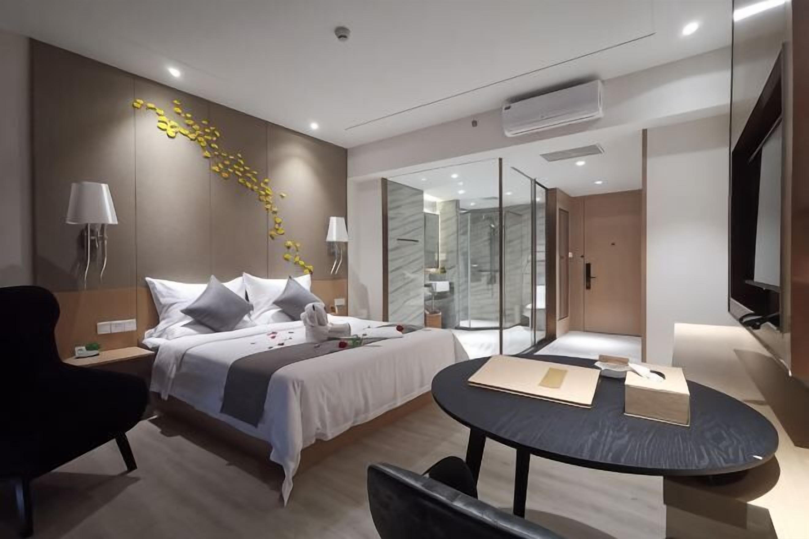 A modern hotel room with a bed, bedside tables, a desk, a chair, a glass shower, and a TV.