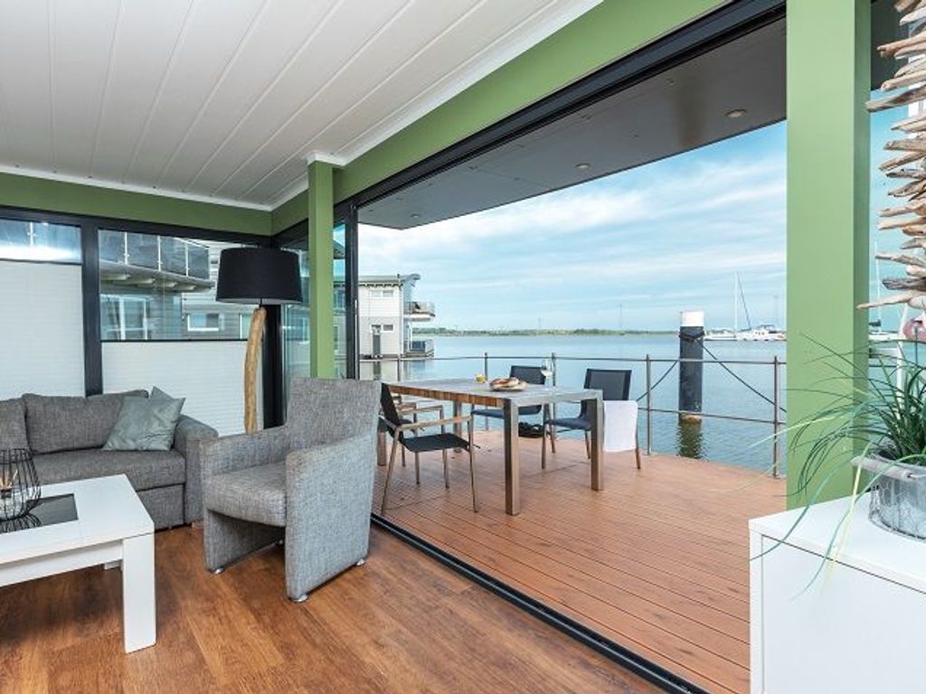 Ein modernes Wohnzimmer mit Holzboden, einem grauen Sofa, einem weißen Couchtisch und einer Essecke mit Tisch und Stühlen. Ein großes Fenster bietet Blick auf das Wasser und Boote.