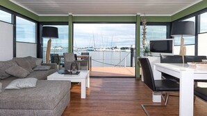 A modern living room with a view of a marina, a white desk, a grey sofa, and a TV.