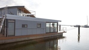 A modern boat with a covered deck and a metal railing, docked at a calm body of water with a clear sky.