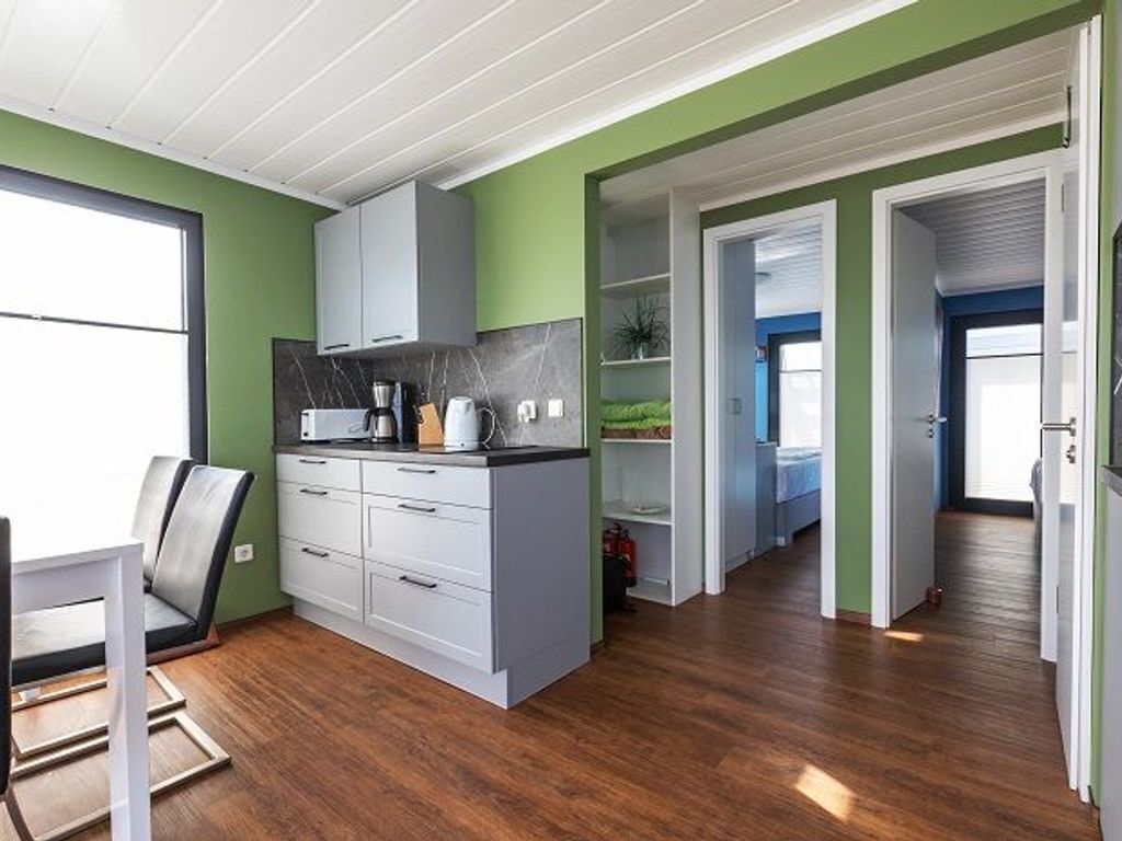 Een moderne keuken met witte kasten, een zwart aanrecht en een raam. Er is een eethoek met een tafel en stoelen. De ruimte heeft groene wanden en een houten vloer.