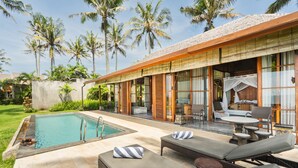 Un espace aménagé au bord de la piscine, avec des chaises longues, une table et des chaises, entouré de palmiers et bordé d’un bâtiment doté de grandes fenêtres.