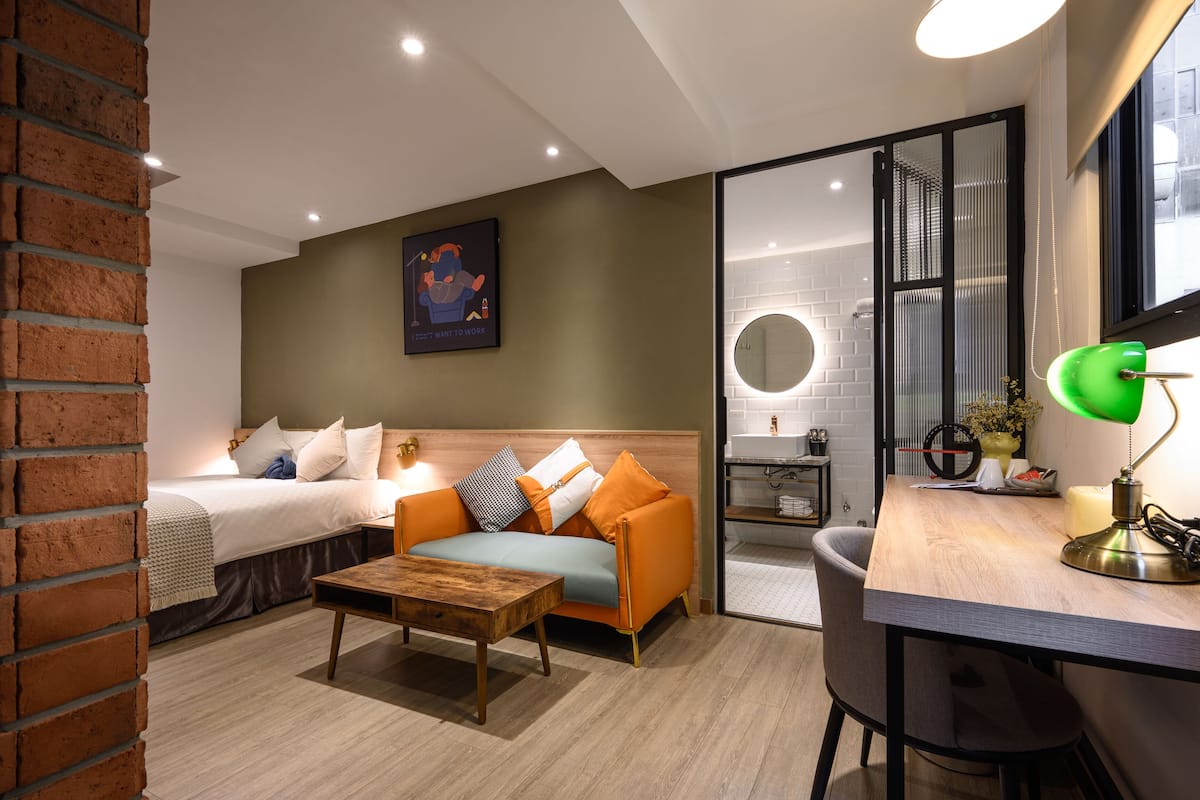 A modern hotel room with a bed, a sofa, a wooden coffee table, a desk with a lamp, and a bathroom visible through a glass door.