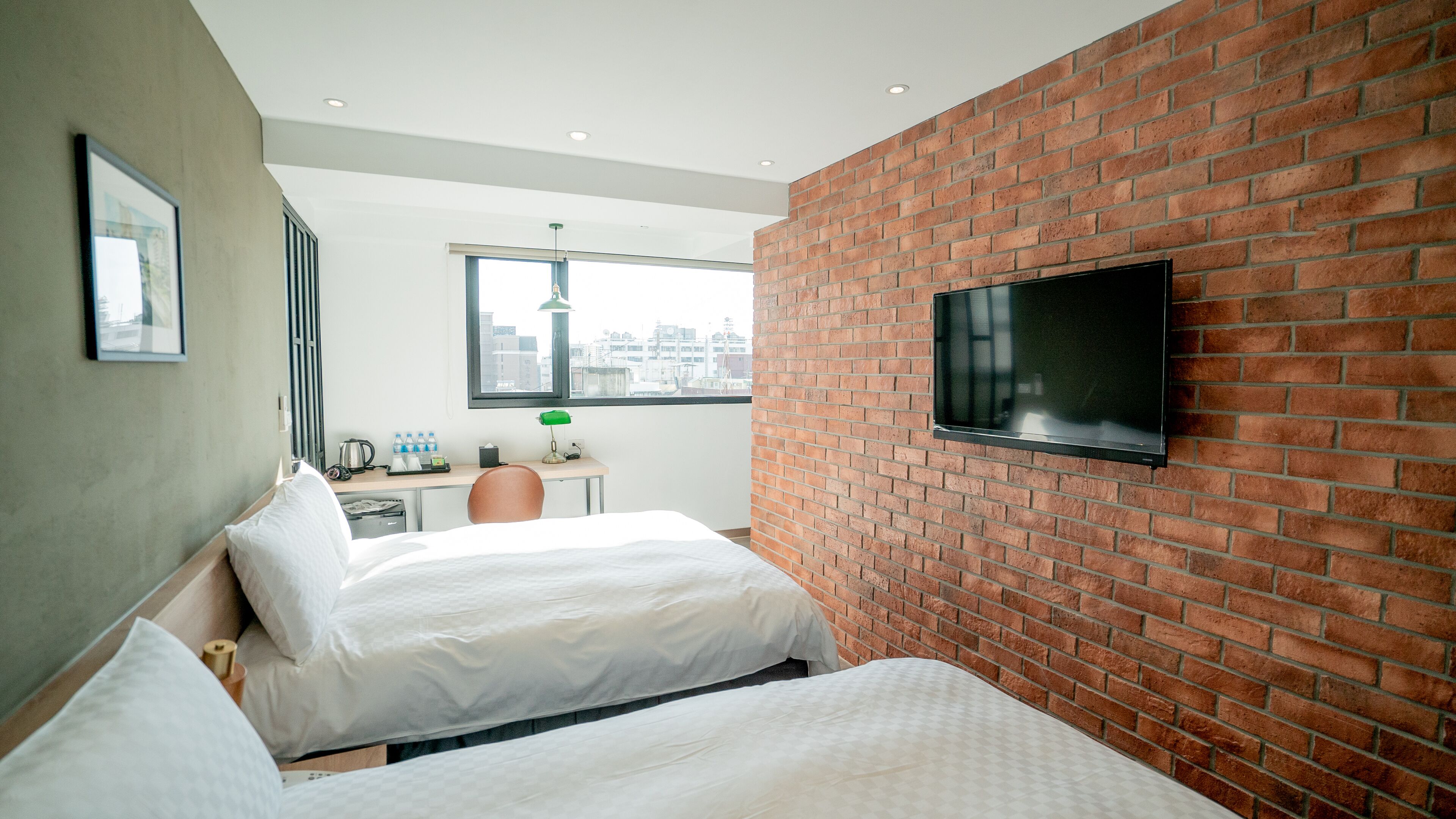Een hotelkamer met twee bedden, een bureau, een aan een bakstenen muur bevestigde televisie en een raam met uitzicht op de stad.