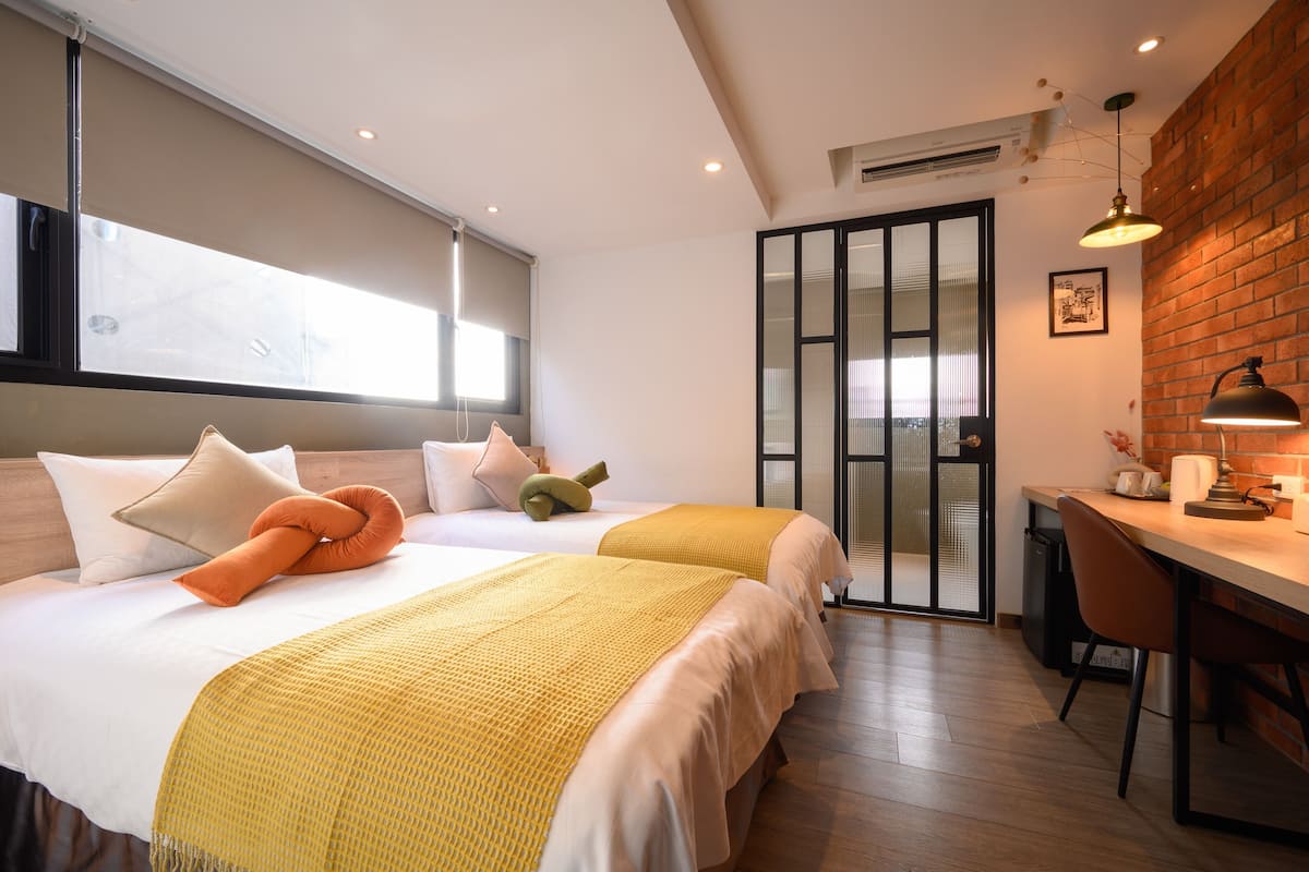 A modern hotel room with two beds, a desk, and a brick accent wall.