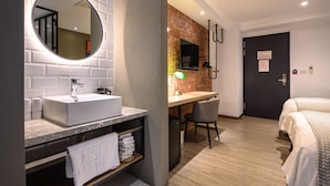 Een moderne badkamer met een witte wastafel, spiegel en handdoekhouder. In de aangrenzende kamer staat een bureau met stoel en een bed.