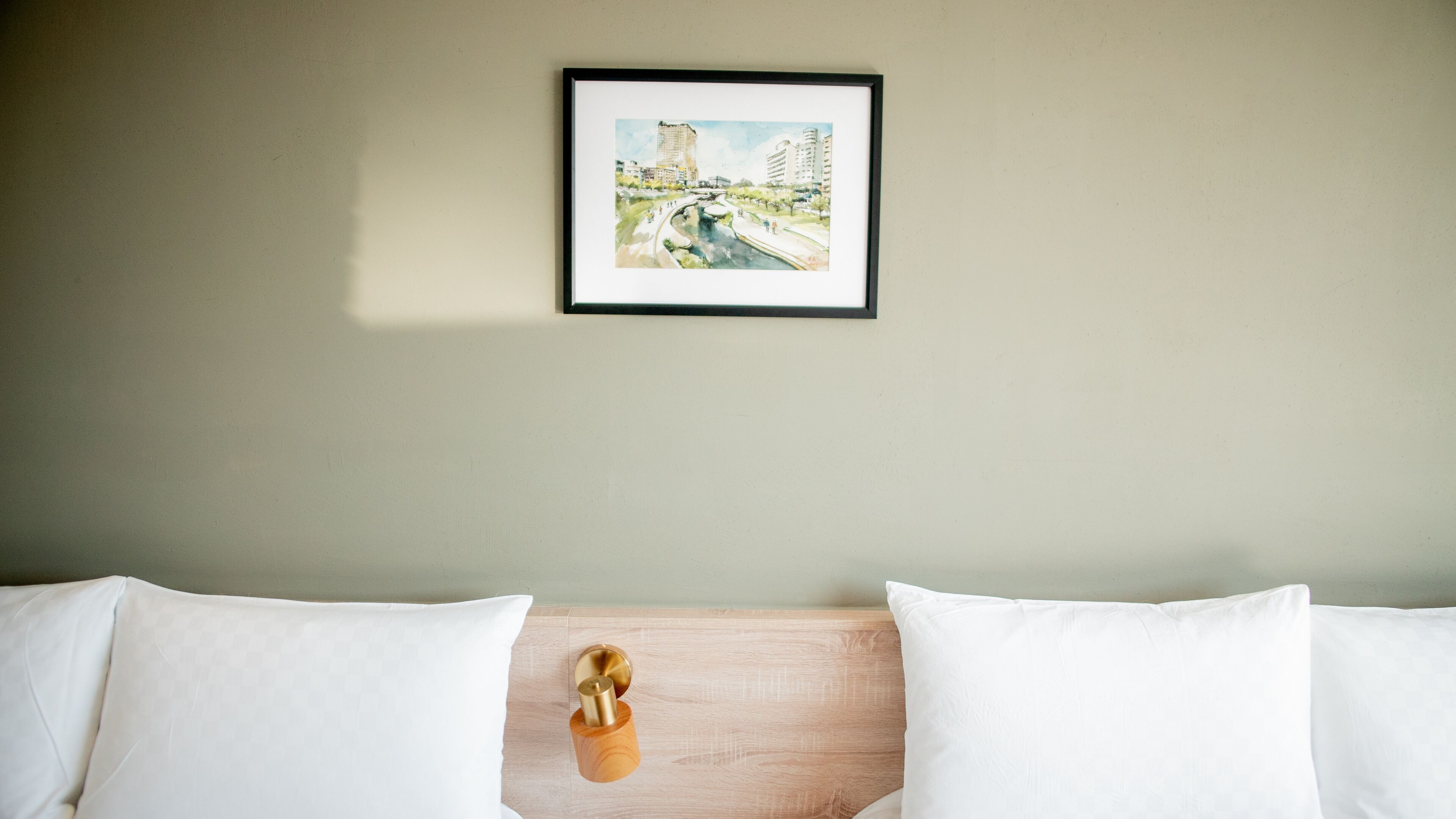 A hotel room with a bed, white pillows, a wooden headboard, and a framed cityscape painting on the wall.