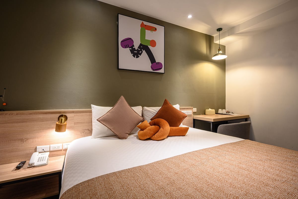 A hotel room with a neatly made bed, a bedside table with a lamp, a phone, and a wall-mounted artwork.