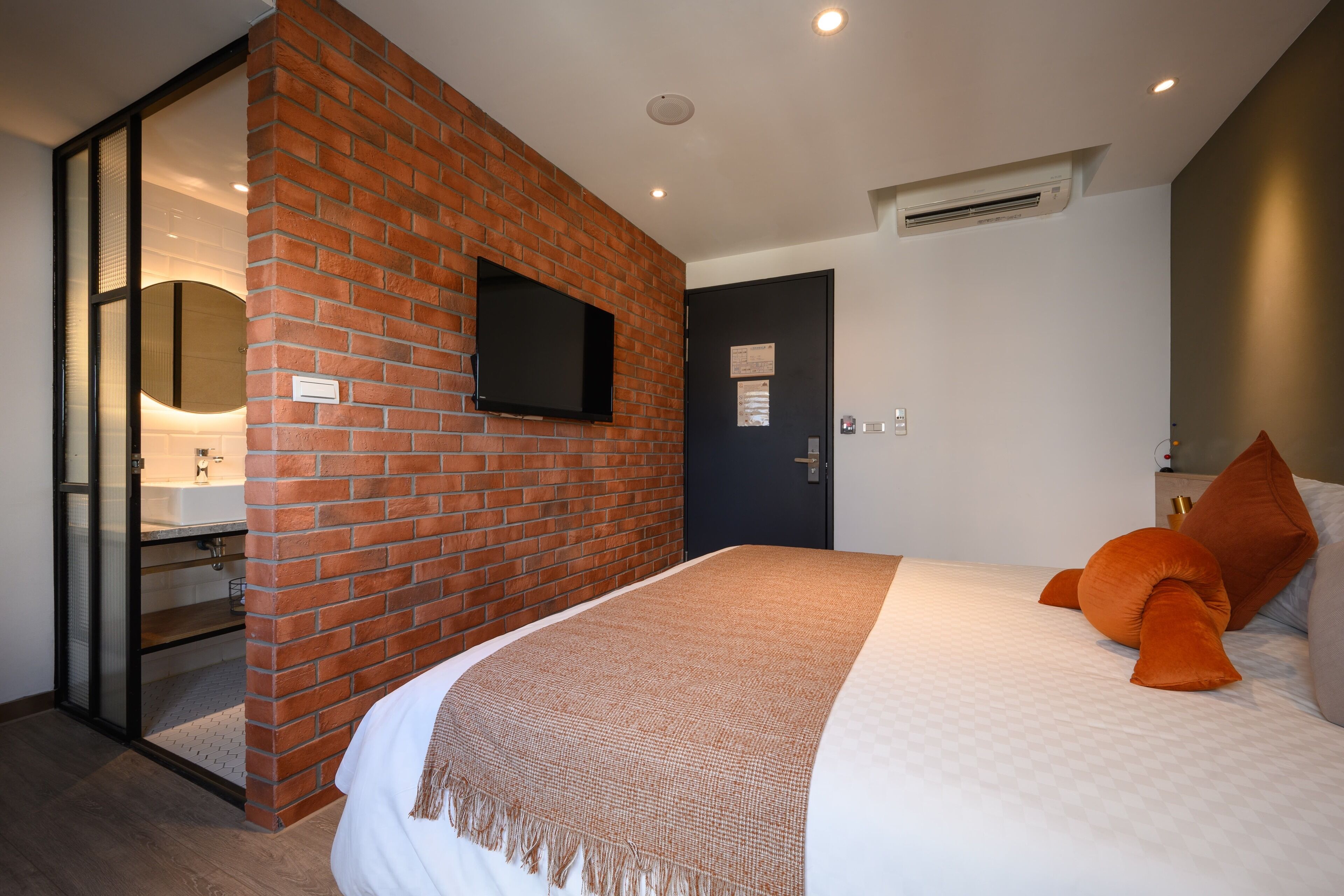 A bedroom with a brick accent wall, a flat-screen TV, a door with a sign, and a bed with orange pillows.