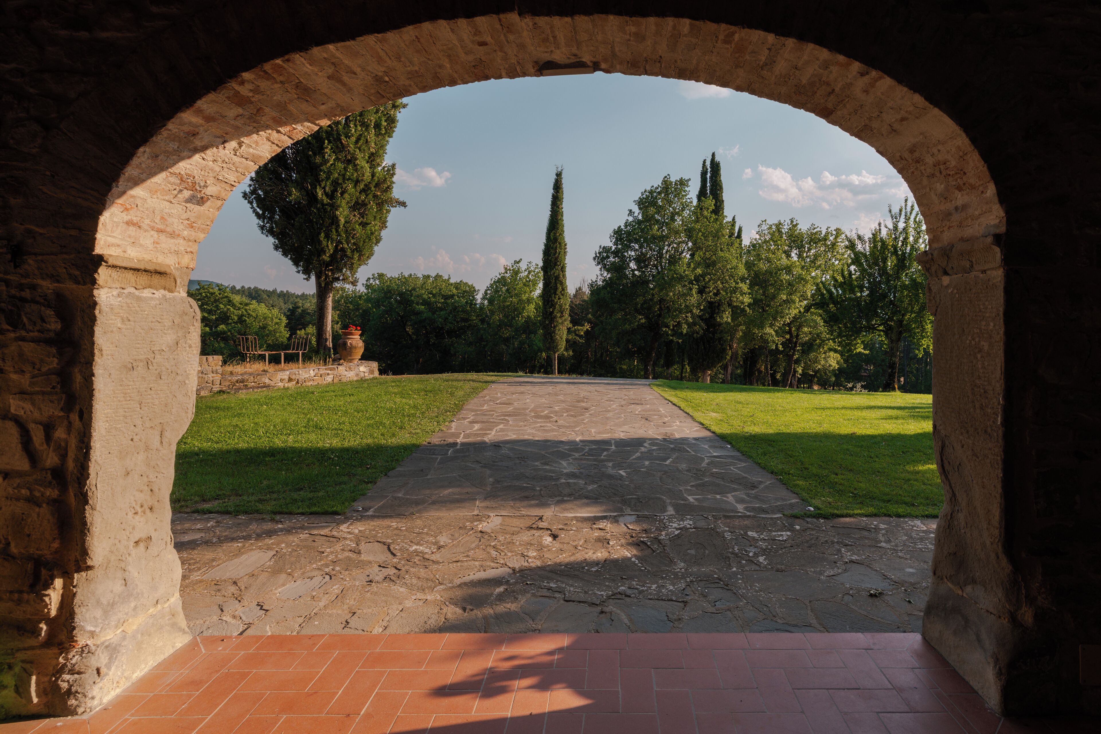 An arched stone pathway leading to a grassy area with trees and a clear sky.