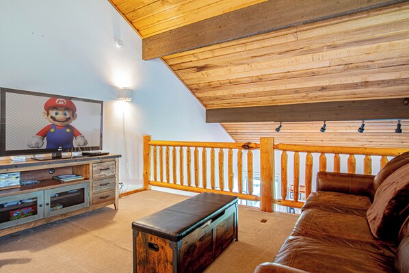 Un salon avec un téléviseur diffusant un jeu vidéo, un plafond en bois, un balcon avec une balustrade et un canapé en cuir.