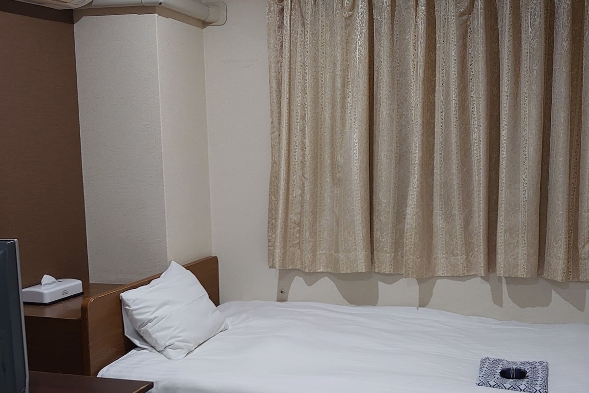 Camera d'albergo con un letto singolo, un comodino con una lampada, un condizionatore d'aria a muro e una finestra con tende a motivi.