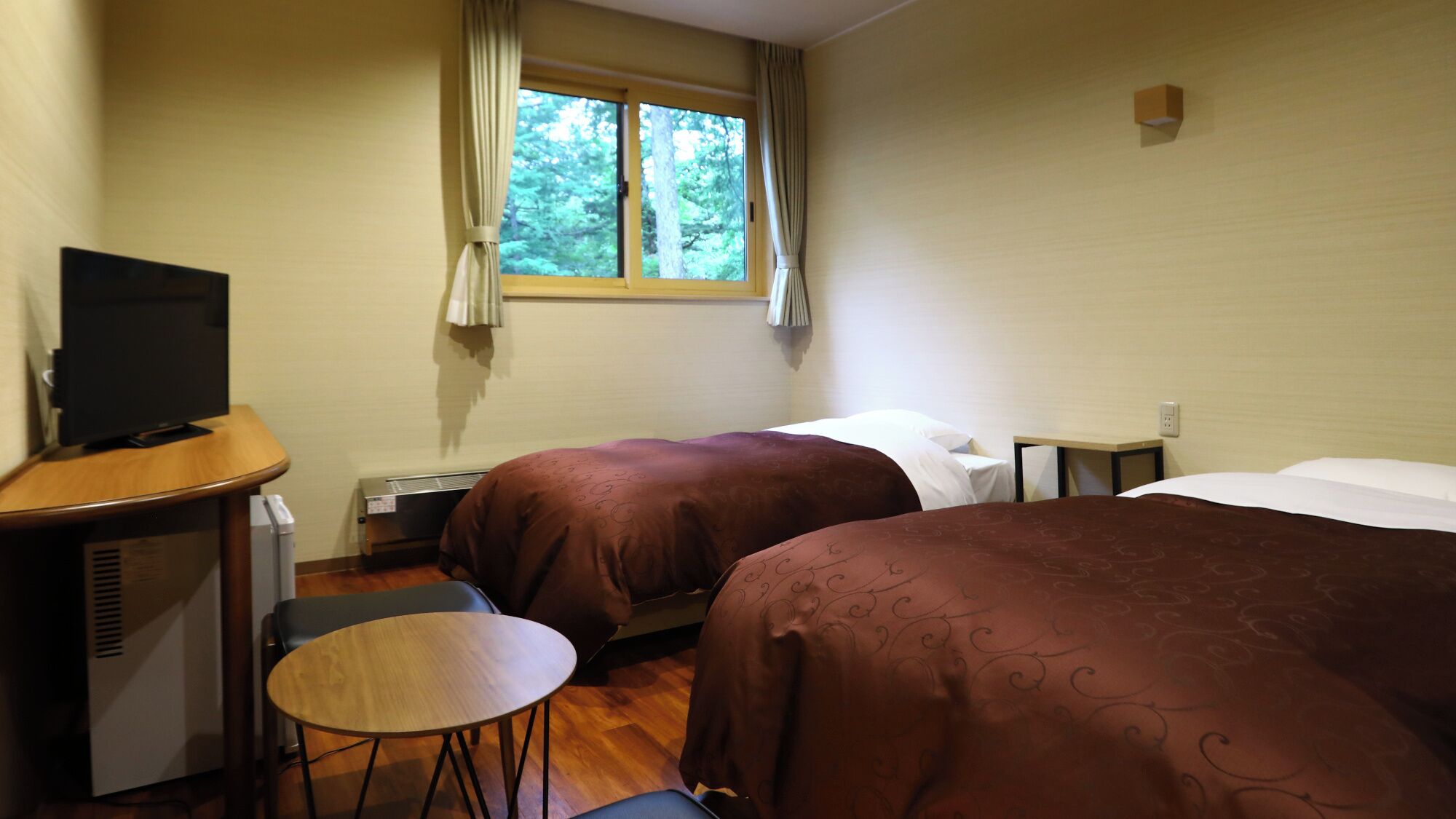 A hotel room with two beds, a desk with a TV, a small table, and a window with curtains.