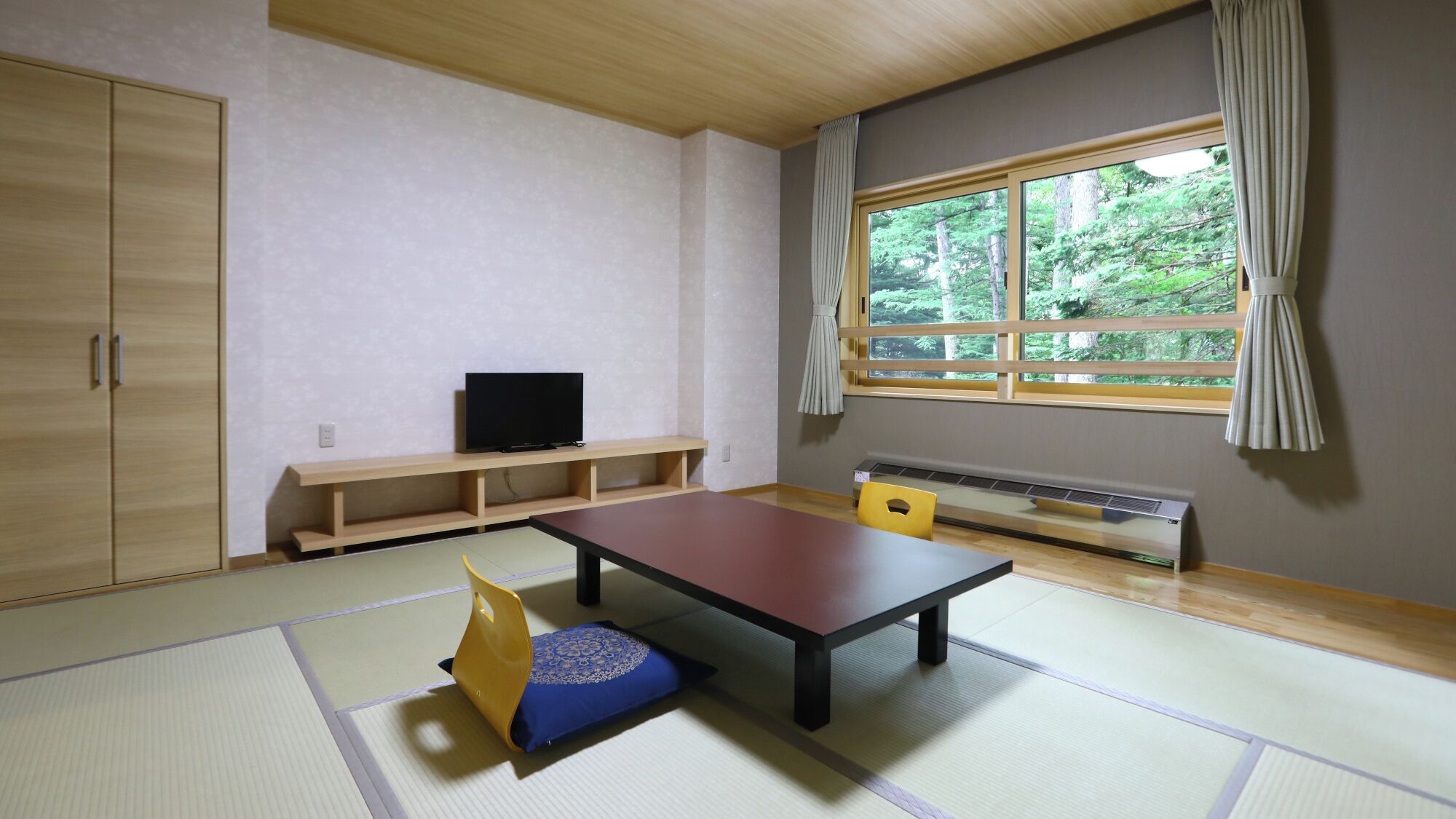 A room with tatami flooring, a low table, a television on a shelf, a window with curtains, and a radiator.