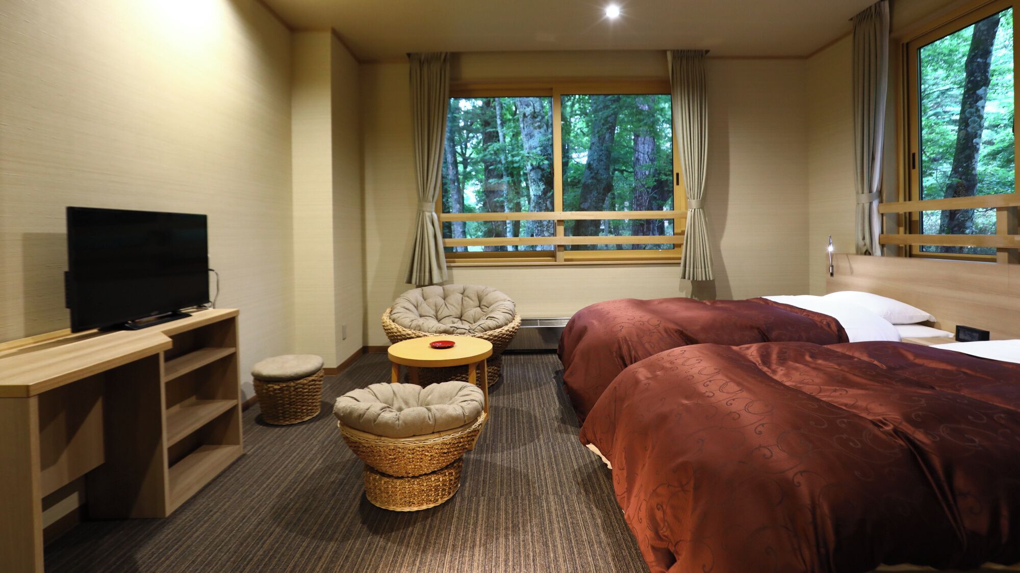 A hotel room with a TV, two beds, a desk, and a window with a forest view.