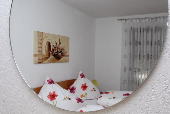 A room with a round mirror reflecting a bed with floral bedding, a painting on the wall, and a window with sheer curtains.
