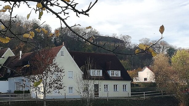 Une maison blanche avec un toit en pente, entourée d’arbres et avec une colline en arrière-plan.