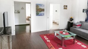 Un salon moderne avec un téléviseur à écran plat, un canapé gris, une table basse en verre et un tapis rouge. Un tableau est accroché au mur et une porte donne accès à une autre pièce.