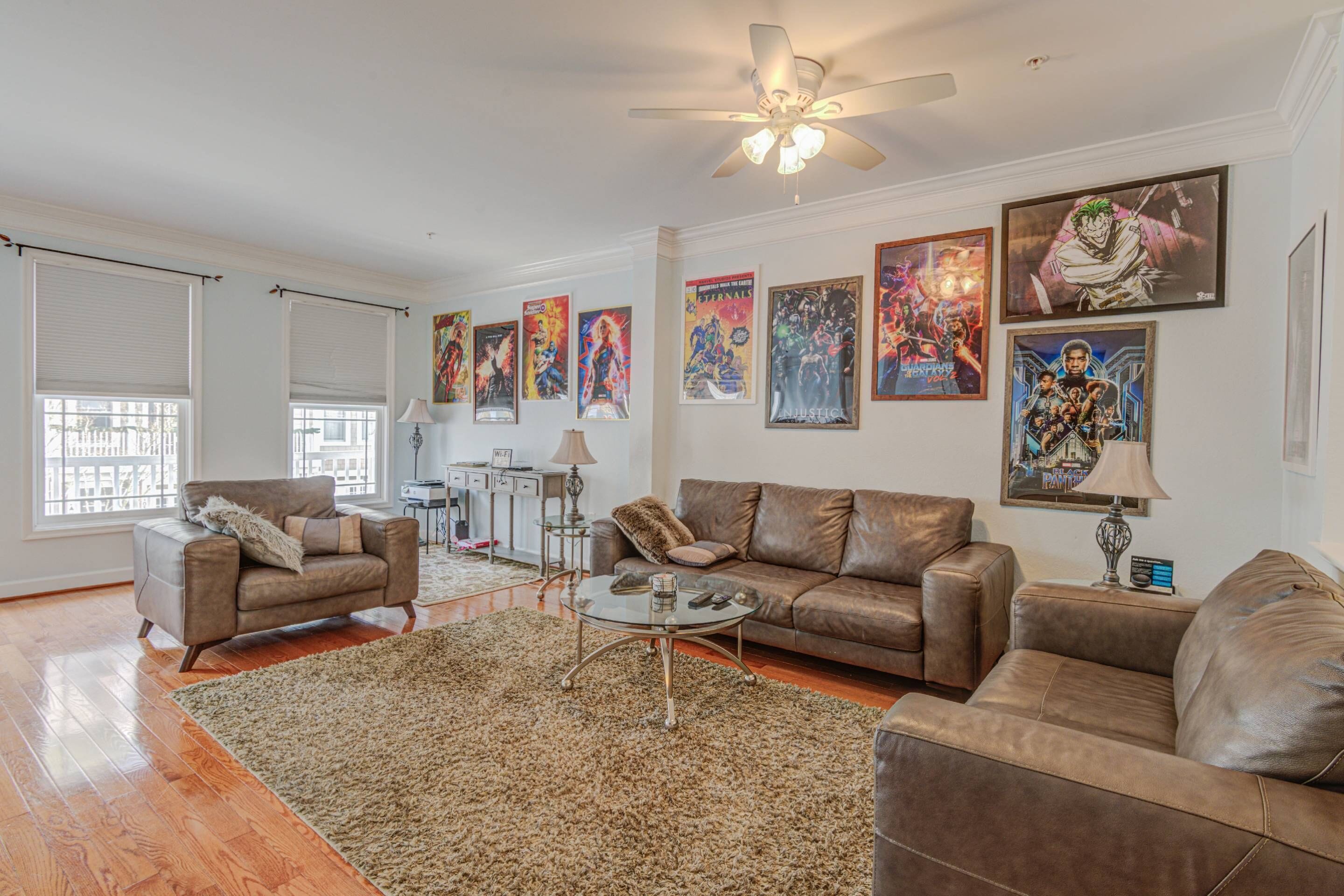 A living room with a brown leather sofa, a glass coffee table, and a large shaggy rug. There are several framed posters on the wall and a ceiling fan above.