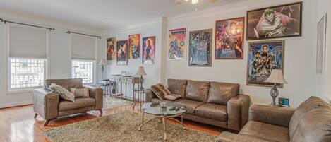 A living room with a brown leather sofa, a glass coffee table, and a large shaggy rug. There are several framed posters on the wall and a ceiling fan above.