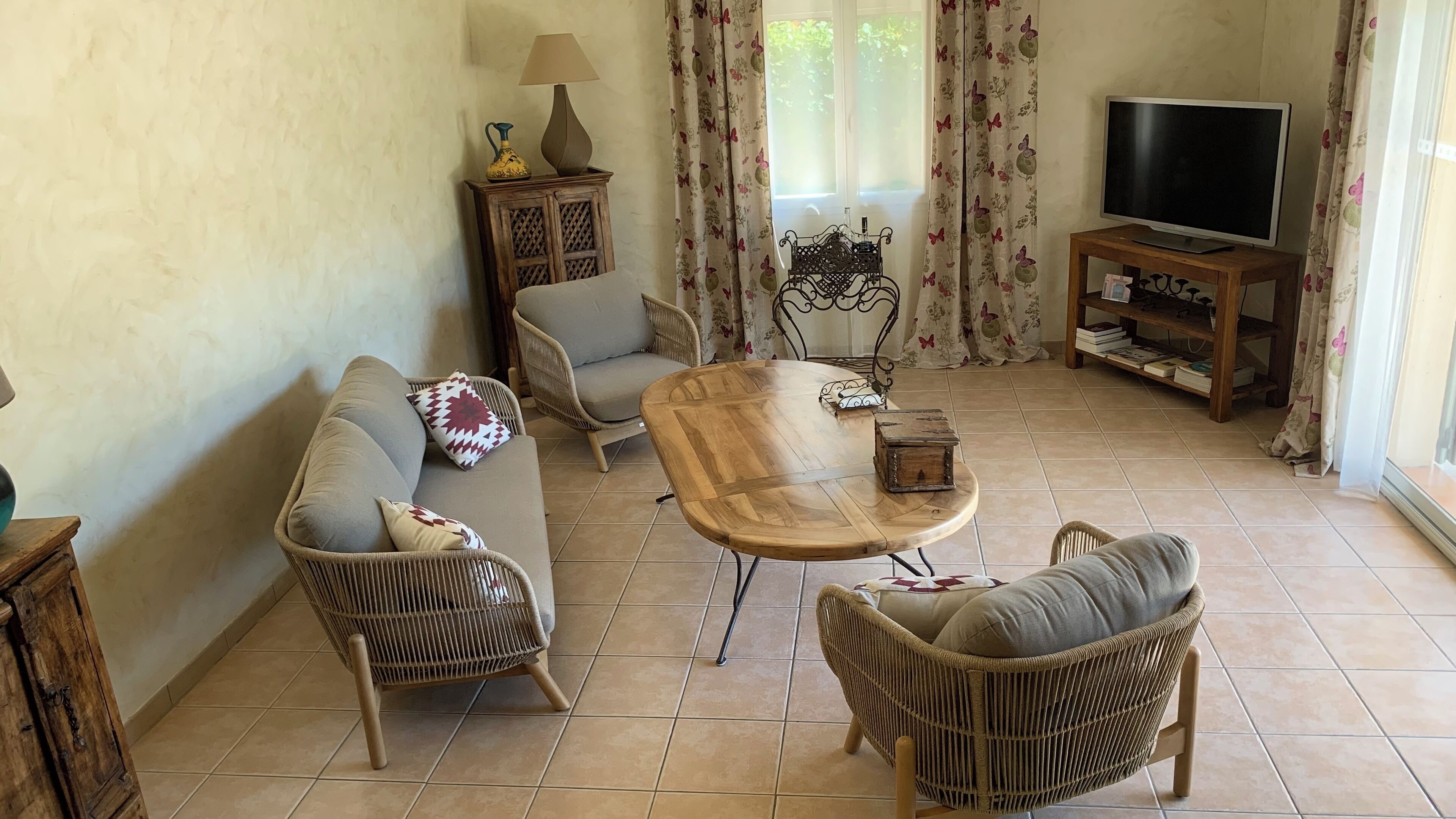 Una sala de estar con un sofá, dos sillones de mimbre, una mesa de centro de madera, un televisor y un aparador con una lámpara.