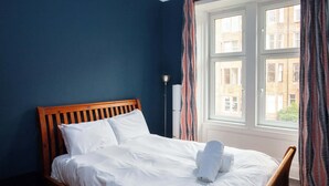 A neatly made bed with white linens and a rolled towel, a wooden headboard, and a window with curtains.
