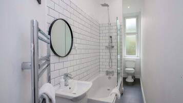 Un baño moderno con pared de azulejos blancos, un espejo redondo, un lavabo, una bañera, una ducha y un inodoro.