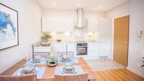 A modern kitchen with white cabinets, a stainless steel range hood, and a dining area with a wooden table set for two. - Stylish 2-Bed Apartment Near Glasgow Green (Glasgow City)