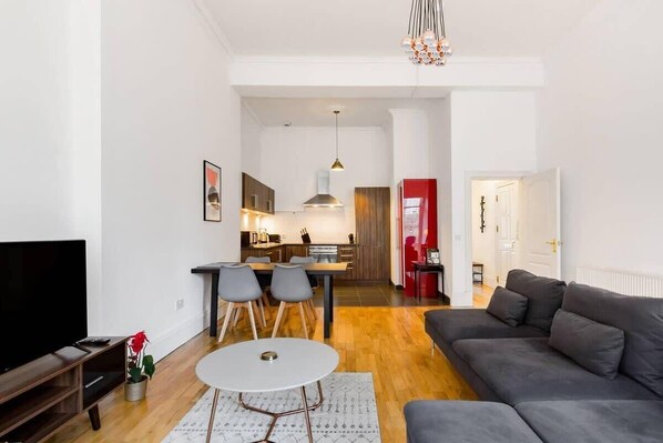 A modern living room with a flat-screen TV, a grey sofa, a dining table with chairs, a kitchen area, and a hallway leading to a door.