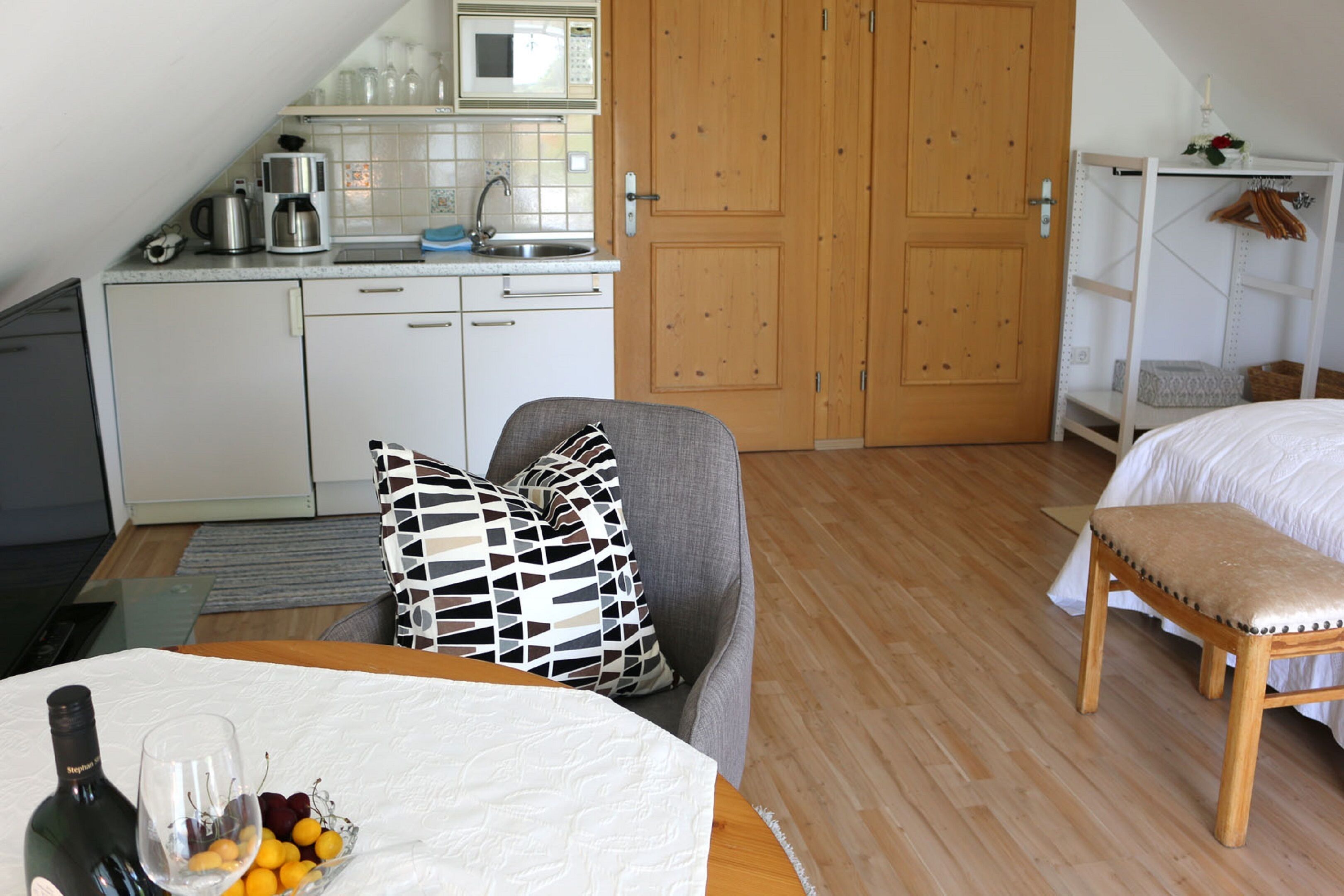 Eine kleine Küche mit Mikrowelle, Kaffeemaschine und Spüle. Eine Holztür und ein Bett mit weißer Bettdecke sind ebenfalls vorhanden.