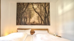 A bed with white linens and a wooden headboard, a nightstand with a lamp, and a large forest-themed painting on the wall. - Peter-Anderl-Hof (Pfreimd)