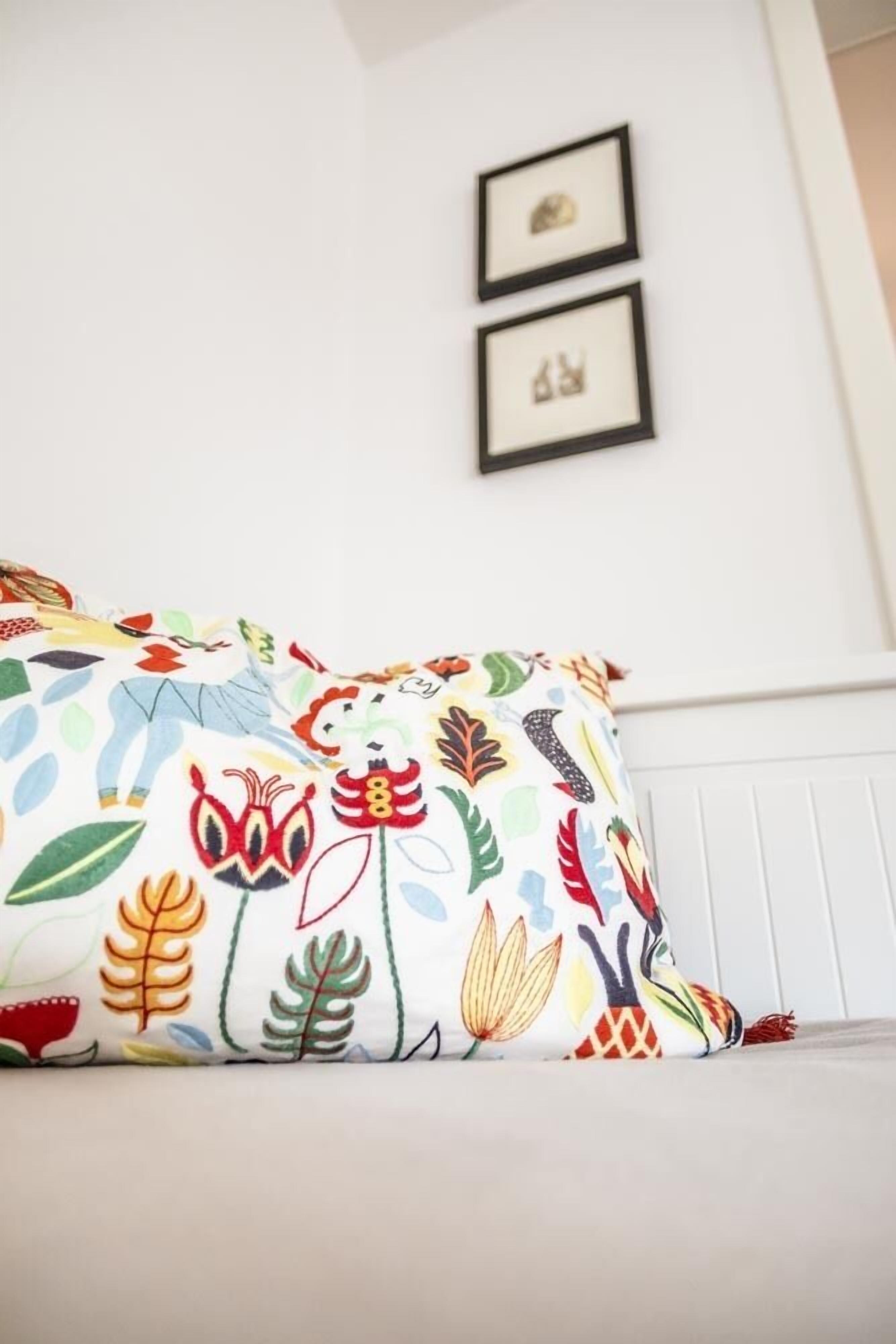 Una cama con un cojín floral de colores y un cabecero blanco. Encima de la cama, hay dos cuadros enmarcados en la pared.