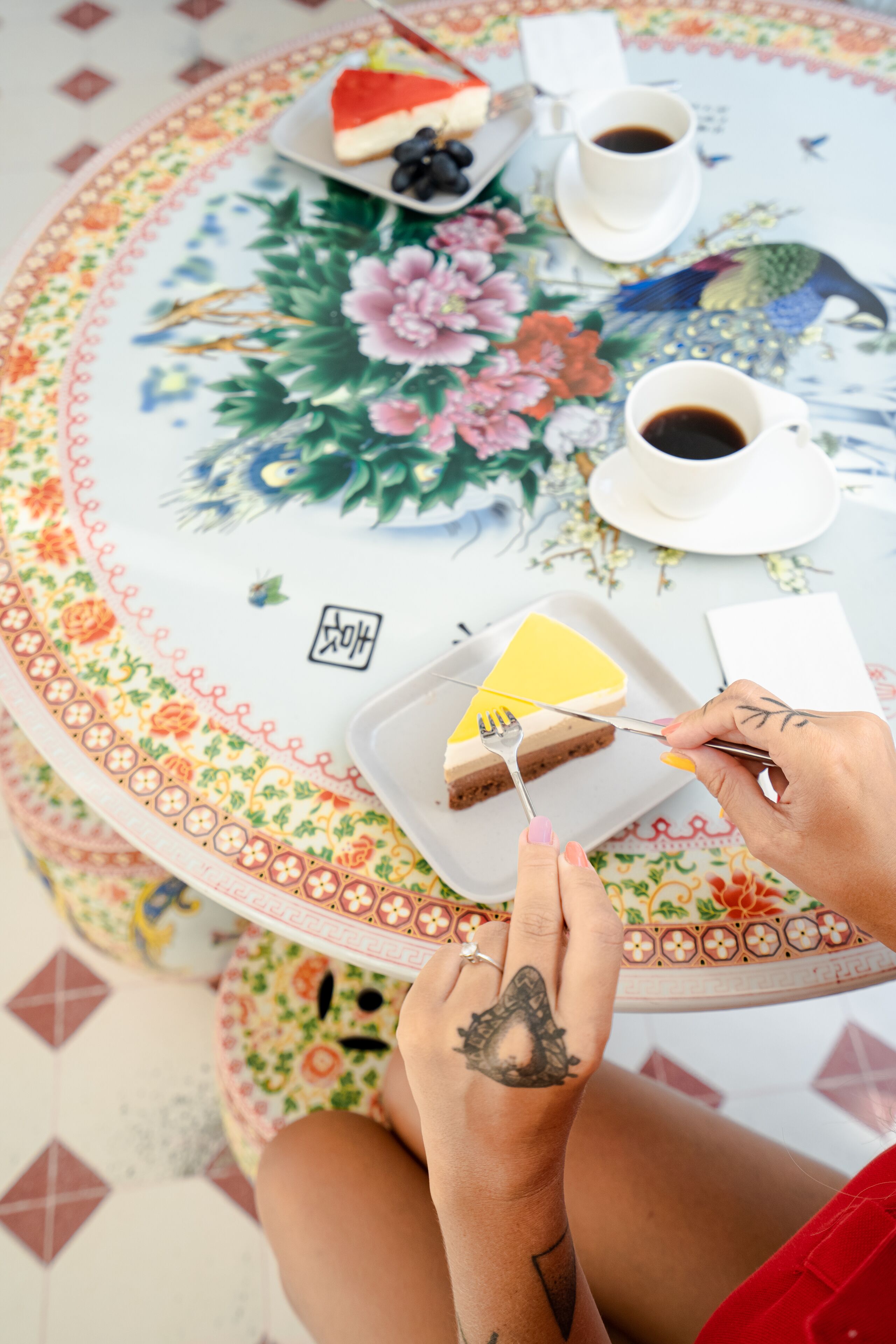 A round table with a floral tablecloth, a plate with a slice of yellow cake, a cup of coffee, and a fork.