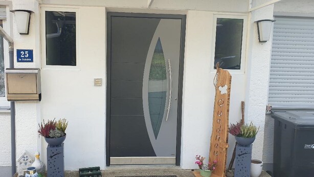 A modern house entrance with a dark-colored door, a small window, and a number '25' sign. There are potted plants and a wooden decorative piece near the door. - Ferienwohnung Quednau (Grafenau)