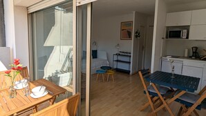 Un appartement moderne doté d’un balcon, d’un coin repas avec des meubles en bois, d’une cuisine avec des placards blancs et d’une vue dégagée sur l’extérieur, visible à travers la porte vitrée.